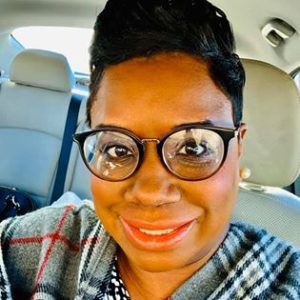 Woman smiling at the camera, from the neck up. She's wearing black rimmed glasses and a plaid shawl.